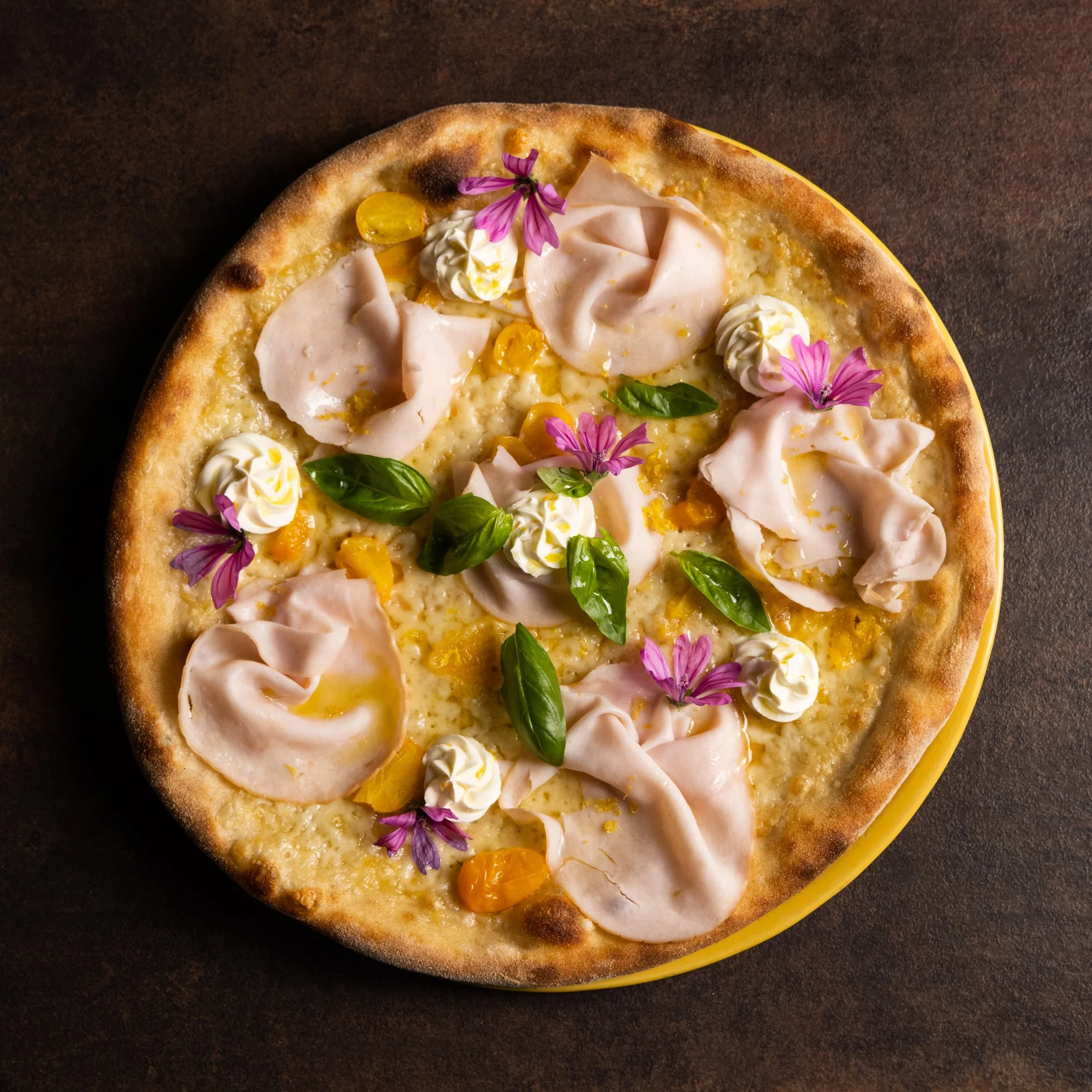 Pizza bianca con mortadella, ciuffi di crema, datterini gialli, basilico e fiori di malva, vista dall'alto